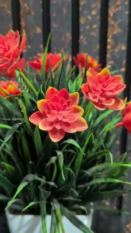 A close-up shot focuses on a potted artificial plant with bright orange and yellow flowers and green leaves. The plant is in a white square pot. The background is blurred, showing vertical dark lines against a textured, golden backdrop. The camera slowly zooms out, revealing more of the plant and its surroundings.