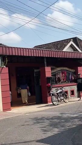 The video shows the exterior of a house with a red facade and a dark-colored roof. The house has an open entrance leading into a dimly lit interior. To the right of the entrance is a black metal gate with ornate gold accents. In front of the house, parked on the paved street, are two motorcycles, one silver and one black. A person wearing a black t-shirt and a black baseball cap stands near the motorcycles. A woman wearing a blue top and a long yellow skirt is walking towards the house entrance, carrying a bag in her right hand. The sky above is visible, with some clouds and power lines.