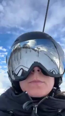 A person wearing a black ski helmet and mirrored goggles looks directly at the camera. The goggles reflect a snowy mountain landscape with ski lifts. The person has pursed lips. They are wearing a black jacket zipped up. The sky is blue with white clouds. A cable from the ski lift runs above the person.