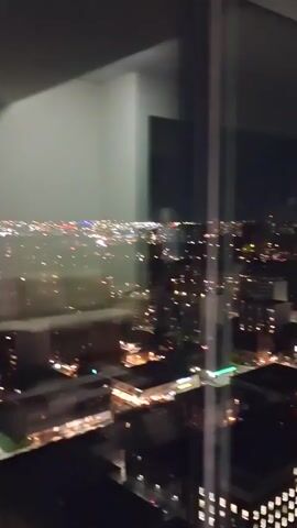 A slow pan across a city at night as seen through a window. The city lights are visible in the distance, with some buildings in the foreground. The reflection of a person and possibly a phone is visible in the window.