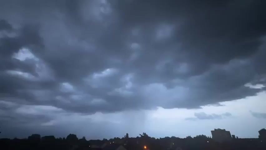 The video depicts a dark, overcast sky with rain falling from a cloud. The silhouette of a town, including buildings and trees, is visible in the foreground.