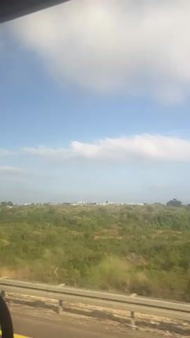 The video shows a view from inside a moving vehicle looking out at a landscape. The landscape consists of rolling hills covered in green vegetation and scattered trees. In the distance, there appears to be a town or settlement with buildings. The sky is blue with scattered white clouds. A roadside barrier is visible in the foreground. The video appears to be recorded during daylight hours.