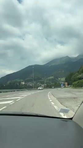 The video shows a view from inside a car traveling on a paved road. The road curves to the right, with a white line marking the lane. On the left side of the road, there is a divided highway with white and yellow lines. In the distance, there are mountains covered in green trees and some clouds. There are also some buildings visible on the side of the mountain. Several cars are seen on the road, and there are streetlights along the highway. There are also road signs visible on the right side of the road. The sky is cloudy. The dashboard of the car is visible at the bottom of the frame.