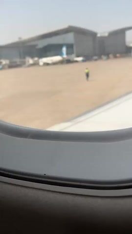 This is a first-person perspective video taken from an airplane window. The scene outside the window shows an airport tarmac under a clear sky. A modern airport terminal building is visible in the background, with large glass windows and a grey, angular roof structure. Several airplanes are parked on the tarmac, including one with a blue and white livery and another with a red and white logo on its tail. An airport worker in a yellow vest and blue pants is standing in the middle of the tarmac. The foreground of the video is dominated by the white wing of the airplane the camera is in, with visible seams and markings on its surface. The wing extends across the lower portion of the frame, partially obscuring the view of the tarmac and terminal. The video captures the view as the airplane presumably taxis or is preparing for takeoff.