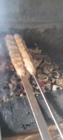 The video shows several pieces of ground meat kofta on skewers cooking over hot coals in a grill. Smoke rises from the grill. The kofta are elongated and appear to be browning and developing grill marks. The environment looks like an outdoor cooking area. There are no people visible in the video.