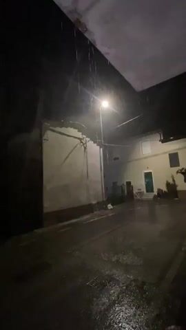 The video shows a street at night during a rainstorm. A street light illuminates the scene, casting long shadows. Water is flowing in the street and the pavement is wet. There are buildings on either side of the street. In the foreground, a person's feet wearing white slides are visible on a wooden porch. Next to the person's feet is a large, clear plastic bag filled with what appears to be trash.