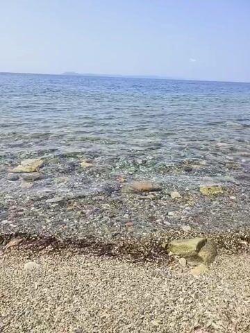 The video shows a slightly blurry, low-resolution view of a pebble beach and clear ocean water under a bright, sunny sky. The water is transparent enough to see the pebbles and rocks beneath the surface. In the distance, a faint landmass or island is visible on the horizon. The camera is positioned low, close to the ground, looking out towards the sea. There are no people or discernible objects other than the natural elements of the beach and ocean.