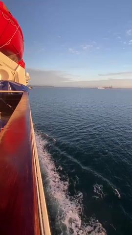 A side view of a large boat moving through the ocean at sunrise or sunset. The wooden railing of the boat is in the foreground on the left. Red life preservers are visible at the top of the frame. The ocean is dark blue and choppy with white foam trails behind the boat. The sky is a pale blue with a few wispy clouds, and the sun is low on the horizon, casting a warm glow. In the distance, a hazy coastline with some structures is visible. The video is shot from a slightly low angle, looking out towards the horizon.