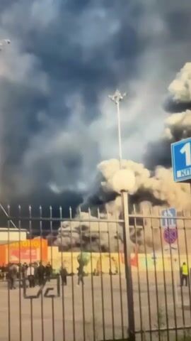 The video captures a large plume of dark smoke billowing into the sky from behind a metal fence. On the fence, a blue sign with the number "1" and some Russian text is visible. In the foreground, several people are standing and observing the smoke. The environment appears to be an industrial or urban area with buildings and streetlights visible in the background. The overall scene suggests a significant fire or industrial incident.