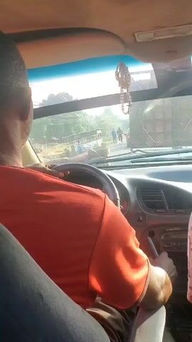 The video shows the perspective of a driver in a car approaching an intersection. The driver, wearing an orange shirt, is seen from the back. The dashboard of the car is visible, along with a beaded ornament hanging from the rearview mirror. Through the windshield, a large truck is parked on the right side of the road, partially blocking the view. Two men are standing near the truck, one dressed in blue. The surrounding environment appears to be a rural or semi-rural area with buildings and power lines visible in the background. The car seems to be slowing down as it approaches the intersection. There is also a partial view of a person in a pink patterned shirt sitting in the passenger seat.