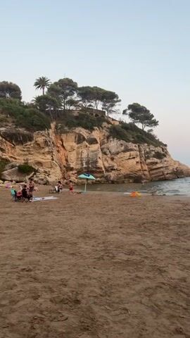 A vertical video shows a beach scene with a sandy shore in the foreground. In the middle ground, there is a body of water with several people swimming and one person on a paddleboard. Further back, a rocky cliff with trees on top juts out into the sea. The sky is clear with a gradient of light blue to pale pink, suggesting it might be late afternoon or early evening. There are a few beach umbrellas and some people sitting on the sand on the left side of the frame. The video pans slightly to the right, showing more of the water and the horizon.