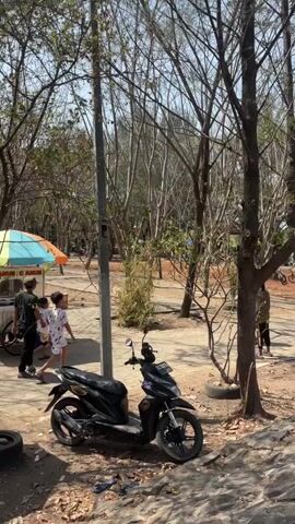 The video shows a sunlit outdoor scene with several people walking in a park-like area with many trees. In the foreground, a black scooter is parked on a dirt path, with fallen leaves scattered around. To the left, a food cart with a colorful umbrella is visible, and two people are standing near it. A tall pole with some wires is also in the foreground. Further into the scene, several individuals are walking on a paved path, with trees and some bamboo plants in the background. The overall atmosphere appears casual and relaxed, possibly a recreational area.