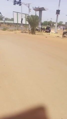 The video shows a street scene on a sunny day. The road is made of sand and dirt, and the sky is a bright, clear blue. Several vehicles are visible, including a yellow three-wheeled auto rickshaw and a black motorcycle. A man wearing a dark shirt and a hat is sitting on the motorcycle.  Other people are visible in the background and standing near the side of the road. Traffic lights stand tall at the intersection. The scene appears to be in a less-developed area, with dirt roads and sparse vegetation.
