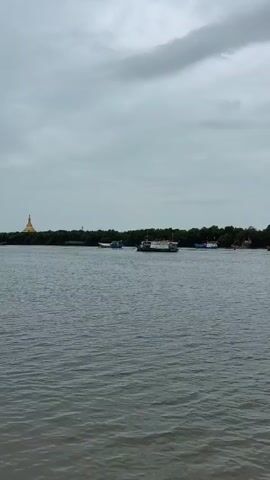 A drone shot pans across a body of water with a treeline and a building in the distance under a cloudy sky. Several boats are visible on the water.