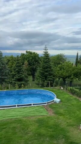 The video shows a backyard with a pool on a cloudy day. The pool is oval-shaped and filled with water, covered by a blue pool cover. To the left of the pool, there is a green tarp on the grass. Behind the pool, there are various trees. Beyond the trees, a city is visible in the distance, under a cloudy sky. The camera moves slightly from right to left, showcasing more of the landscape. The grass is green and well-maintained.
