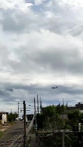 A distant airplane flies through a cloudy sky over a neighborhood street. The street is lined with houses, power lines, and trees. A sign saying "Casas de Venta" is visible in the bottom right corner of the frame. A person walks down the street.