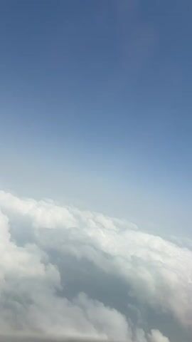 The video depicts an aerial view of clouds from an airplane window. Above the clouds is a clear blue sky. The airplane window frame is visible in the lower right corner. The view is mostly still. The lighting is bright.