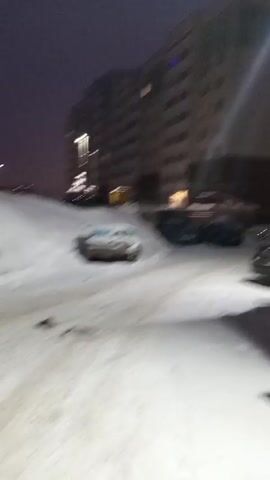 The video shows a snowy, urban environment at night. There are apartment buildings, cars parked on the street covered in snow, trees without leaves, and a supermarket sign that reads "МАГНИТ". The camera moves, panning across the scene. There are some streetlights, and lights are on in some of the buildings.