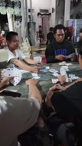 The video shows a group of men sitting around a wooden table playing cards. There are at least four men visible. One man on the left is wearing a white t-shirt and has tattoos on his arm. He is holding a fan of cards. Another man in the center is wearing a black hoodie with "DUKE" written on it. He is also holding cards. The table is covered with playing cards and some money. The background appears to be outdoors, possibly a yard or patio, with some decorations and other people in the distance. The lighting suggests it is nighttime or dusk. The overall atmosphere appears to be casual and social.