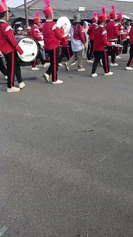 The video shows a marching band in a street parade. The band members are wearing red jackets with black trim, black pants, and red hats with pink plumes. Some members are playing drums, while others are holding flags. The parade is taking place in a populated area with onlookers on both sides of the street. There are also motorcycles parked along the side of the road. The atmosphere appears festive.