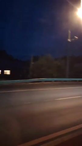 The video depicts a nighttime highway scene, viewed from the perspective of a moving vehicle. The sky is dark blue, with a few streetlights visible, casting a yellowish glow. Trees line the side of the highway, their forms blurred due to the motion. The road itself is asphalt, with yellow lane markings that are also streaked with light. Several vehicles are seen passing by, their headlights and taillights creating streaks of light. One vehicle appears to be a red bus, while another is a white car. The overall impression is one of speed and the blur of a night drive.