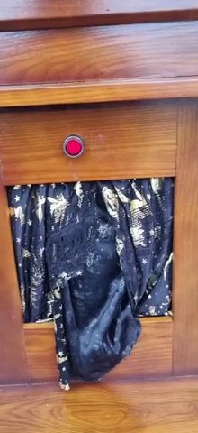 A hand enters the frame from the left and presses a red button on a wooden surface. The button is located above a black and gold patterned fabric. The fabric appears to be draped or hanging in a recessed area. The hand is partially visible as it interacts with the button.