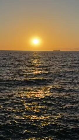 The video shows a wide shot of the ocean at sunset. The sun is a bright, glowing orb near the horizon, casting a golden path across the rippling water. In the distance, a large ship is visible, silhouetted against the warm glow of the sky. The overall scene is tranquil and picturesque, evoking a sense of peace and natural beauty. The camera is static, with no visible movement or interaction. The environment is purely natural, with the focus on the seascape.