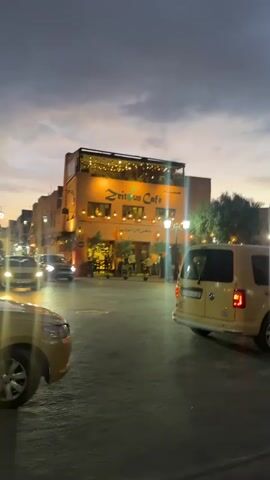 The video shows a street scene at dusk with the Zeitoun Cafe visible in the background. The building is lit up with lights. Several vehicles are present, including a yellow van in the foreground and other cars driving on the street. A person walks across the street in front of the van. A motorcycle passes by. The sky is cloudy.