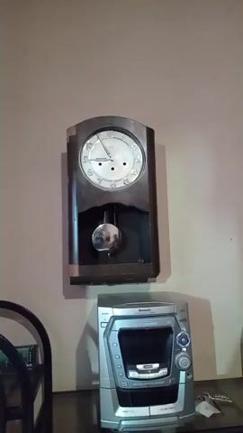 This is a vertical video recording of a pendulum clock mounted on a wall. Below the clock sits a silver stereo system. The clock face displays roman numerals and has three hands. The pendulum is visible swinging back and forth behind a glass panel. The stereo system has a CD player and various buttons and knobs. The background is a plain, light-colored wall. The lighting appears to be artificial. There is no sound in the video.