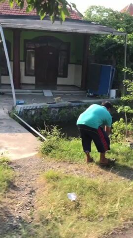 A person wearing a teal shirt and dark pants with red stripes bends down to pick up trash near a ditch. The person appears to be in a yard with a house in the background. The ground is covered with dirt and grass. Sunlight is visible.