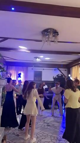 The video shows a group of people dancing in a room. The room has a tiled floor and a wooden ceiling with a chandelier. There are also curtains, plants, and what appears to be a fireplace. Some of the people are dressed in formal attire, while others are in more casual clothing. The lighting in the room changes throughout the video, with some sections appearing to be illuminated by colored lights. The people are engaged in various dance moves, and some are interacting with each other. There is also a piano in the background.