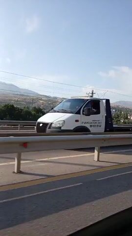 This video captures a roadside view on a sunny day, likely in Charvak, Uzbekistan. The scene is dominated by a highway with a metal guardrail separating lanes. On the highway, a white minivan with a roof rack is moving away from the camera. Behind the guardrail, a train track runs parallel to the road, with overhead power lines supported by utility poles. In the background, a town with houses and trees is nestled against rolling hills. The sky is clear and blue with a few scattered clouds, suggesting fair weather. The sun appears to be high in the sky, indicating it's likely midday. No visible text is present in the video. The overall atmosphere is peaceful and typical of a journey through a semi-rural landscape.