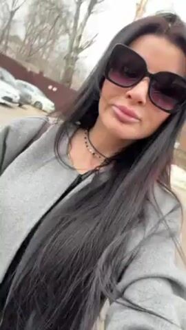 A woman with long black hair is walking down a street. She is wearing large, dark sunglasses and a gray coat. She has a small necklace on. In the background, there are cars, buildings, and trees. The weather appears to be overcast. The woman is holding the camera and filming herself as she walks.