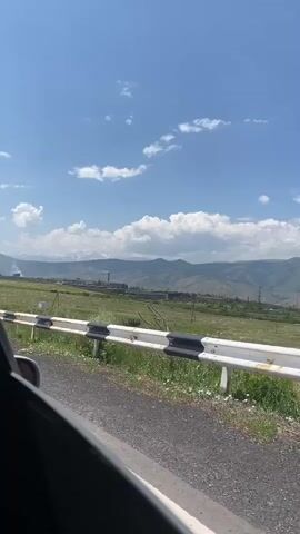 The video is a first-person perspective of someone traveling in a vehicle. The view is out the side window, showing a roadside guardrail with alternating black and white reflective stripes. Beyond the guardrail is a grassy area with some low-lying vegetation and a dirt road. In the distance, there is a valley with rolling hills and mountains. A cluster of buildings, possibly a town or industrial area, can be seen in the valley, along with what appears to be industrial smokestacks. The sky is blue with scattered white clouds. The lighting suggests it is daytime, likely a sunny day. There are no people visible in the video, and no specific actions or interactions are occurring, other than the movement of the vehicle, indicated by the slight blur and changing perspective of the scenery.