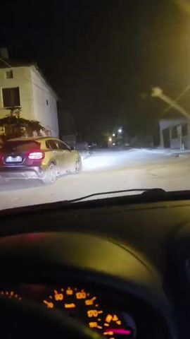A dark-colored Mercedes-Benz GLA with license plate B 7099 XA is seen at night. The car is dusty and appears to be parked or slowly moving on a paved surface. The headlights of another vehicle are visible in the foreground, suggesting this footage is from inside a car. The environment is dimly lit with streetlights visible in the background, illuminating some buildings and trash bins. The rear taillights of the Mercedes are illuminated.