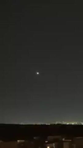 A short, static video shows a night sky. In the center is a single bright star-like light. Below, a dark treeline is visible, with the lights of a distant city just above it. In the foreground, rooftops of buildings can be seen.