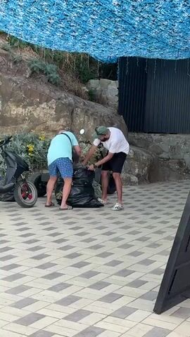 Two men are standing next to a pile of large black trash bags on a patterned tiled surface. A scooter is parked to the left of the men. The man on the left is wearing a light blue t-shirt and blue patterned shorts. The man on the right is wearing a white t-shirt, black shorts, and a green baseball cap. Both men are interacting with the trash bags, with the man on the right appearing to be placing something into one of the bags. Behind the men is a rocky incline covered with some vegetation, and a building with dark siding. A blue mesh covering is suspended above the scene.