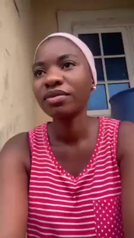 A woman with dark skin and dark eyes is facing the camera, wearing a pink headband and a red and white striped sleeveless top. She is in front of a beige wall with a window behind her to the right. A blue plastic tub is partially visible in the background as well. The woman appears to be speaking, as her lips are moving.