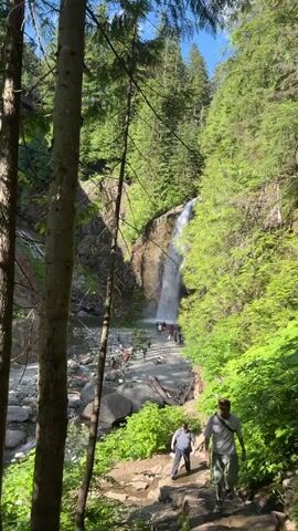 A tall waterfall cascades down a rocky cliff face amidst a lush, green forest. Several people are visible at the base of the waterfall, some standing on the rocky shore and others near the water's edge. A dirt path winds through the forest, with a few individuals walking along it. The sun shines brightly through the trees, illuminating the scene.