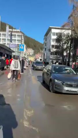 The video shows a street scene on a sunny day. There are people walking on a sidewalk and cars driving on a wet, slushy street. A dark gray BMW SUV with a roof rack is driving towards the camera, followed by other vehicles. There is a pedestrian crossing sign. Buildings are visible in the background, along with a hillside covered with trees.