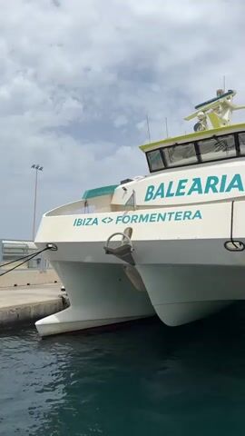 The video shows the front of a white catamaran ferry with "BALEARIA" written on the side in blue letters. Below that, it says "IBIZA <> FORMENTERA". The ferry is docked in clear green water under a cloudy sky. Another ferry, red and white, is visible in the distance to the right. The camera pans slightly to the right, revealing more of the docked ferry and the water.