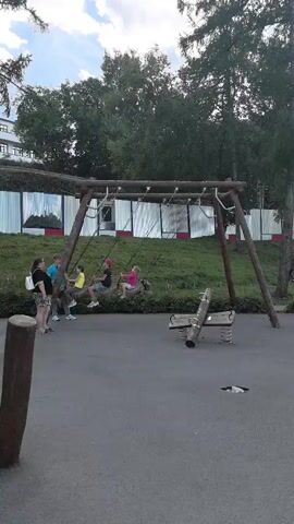 A group of children are playing on a swingset in a park. The swingset is made of wood and has two swings. One child is swinging on the left swing, and two children are swinging on the right swing. A woman is standing next to the swingset, watching the children. In the background, there are trees and a fence. To the right of the swingset, there is a seesaw and a merry-go-round. Several other people are visible in the background, sitting on benches or walking. The ground is paved with asphalt.