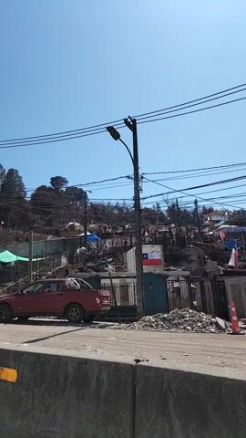 The video shows a roadside view of a landscape affected by a fire. The area appears hilly and is populated with structures, some of which are damaged or destroyed. Chilean flags are visible. A street lamp stands next to utility wires. Cars and trucks are parked along the road, with the camera briefly focusing on a red truck and a white truck. The sky is blue.