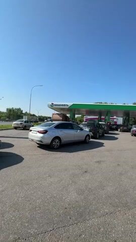 A video captured from inside a vehicle shows a queue of cars at a gas station under a clear blue sky. The gas station has a green and white canopy with the Bashneft logo visible. Several cars are lined up at the pumps, with more cars waiting behind them. The road surface appears to be asphalt. Trees and vegetation can be seen in the background, along with a bright, sunny day. The video pans slightly, offering a view of the different vehicles and the overall scene at the gas station.