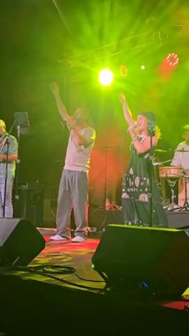 A live music performance is captured on stage under bright green and red stage lights. The stage is equipped with speakers and lighting trusses. Two vocalists, a man and a woman, are prominently featured. The man, wearing a light grey t-shirt and loose grey pants, stands to the left, holding a microphone and facing the audience. The woman, dressed in a long black dress with a celestial pattern and a black headscarf, stands to the right, also holding a microphone and singing. Behind them, other band members are partially visible, including a guitarist on the far left wearing a cap and a patterned shirt, and a percussionist on the far right. The overall atmosphere suggests an energetic concert.