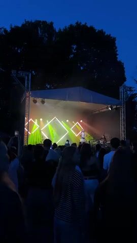 A vertical video captures a live music performance outdoors at dusk. A stage is set up with a white canopy and a backdrop featuring geometric lighting designs in green and pink. A lone performer stands on stage, singing into a microphone. A band is visible behind the singer. The stage is illuminated by bright spotlights. In the foreground, a crowd of people are gathered, facing the stage. The sky above is a deep blue, and the silhouettes of trees frame the top of the scene. The overall mood is energetic and celebratory, characteristic of an outdoor concert.