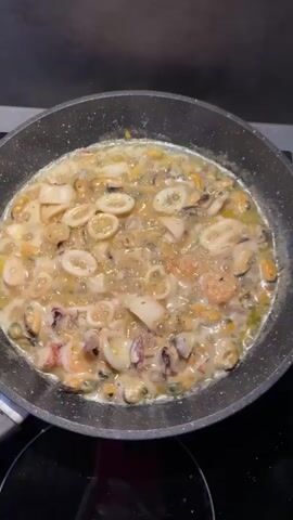 The video shows a kitchen scene. The first shot is a close-up of a frying pan on a stovetop filled with seafood and sauce, with the ingredients simmering. The camera then pans to a table with a glass lid and a spoon. Next, it shows a bowl of cooked pasta, two packages of butter, and other kitchen items on a counter. The final shots are wider views of the kitchen, showing the pasta in the bowl, the window with curtains, and a bottle of dish soap. The overall scene suggests food preparation and cooking. There are no people visible in the video.
