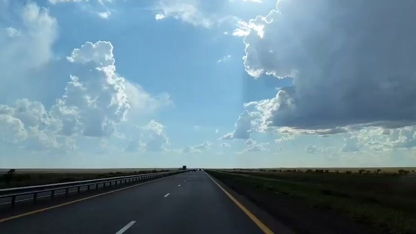 A wide-angle shot from a moving vehicle shows a flat landscape with a two-lane asphalt highway extending into the distance. The sun is bright, peeking through dramatic, fluffy white clouds against a bright blue sky. The highway has white dashed lines dividing the lanes and a yellow line marking the edge. On the left, a metal guardrail runs along the side of the road, and a grassy shoulder is visible. On the right, a field of green grass stretches to the horizon. In the far distance, several vehicles, including what appears to be a truck, are visible on the highway.