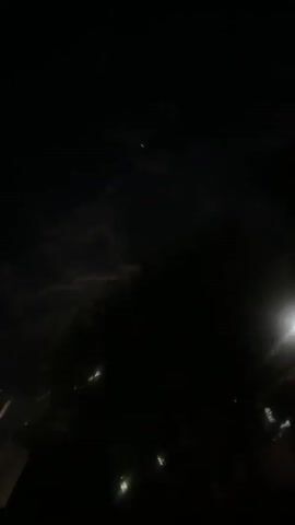 The video shows a night scene, predominantly dark with patches of cloudy sky and what appears to be the moon shining through the gaps.  The camera angle is directed upward.  Below the sky, there's what looks like the silhouette of foliage, possibly trees. Some faint light points, possibly street lamps or lights from buildings, are visible through the leaves. There is movement as the camera is briefly obscured by what appears to be a hand holding a piece of paper or card in front of the lens.