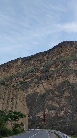The video showcases a scenic drive along a winding road, with a dramatic mountainous landscape dominating the view. The camera pans across imposing, sheer rock faces, revealing layers of sedimentary rock in shades of brown and tan, interspersed with patches of green vegetation clinging to the slopes. A fast-flowing river runs along the base of the mountains, its surface a turbulent green.

A lone tree with lush green foliage stands on the left side of the road, providing a stark contrast to the barren rock. The road itself is paved and appears to be a two-lane highway, with a white barrier running along the edge. In the distance, a utility pole is visible, suggesting some level of infrastructure in this remote area.

The sky is partly cloudy, with wispy white clouds streaking across a pale blue expanse, indicating it is likely daytime, perhaps late afternoon or early morning given the soft light. The overall atmosphere is one of natural grandeur and a sense of vastness. The focus is on the impressive geological formations and the serene beauty of the natural environment. There are no people or animals visible in the footage, and no text is present. The scene conveys a feeling of peaceful exploration and appreciation for rugged natural landscapes.